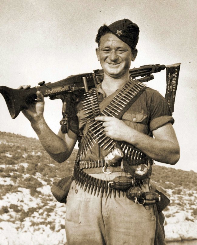 Figure 1 — A Yugoslav partisan on the island of Vis in August 1944 with a captured MG 42 on his shoulder and belts of ammunition across his chest; according to the photograph's description, the flag in the mu…