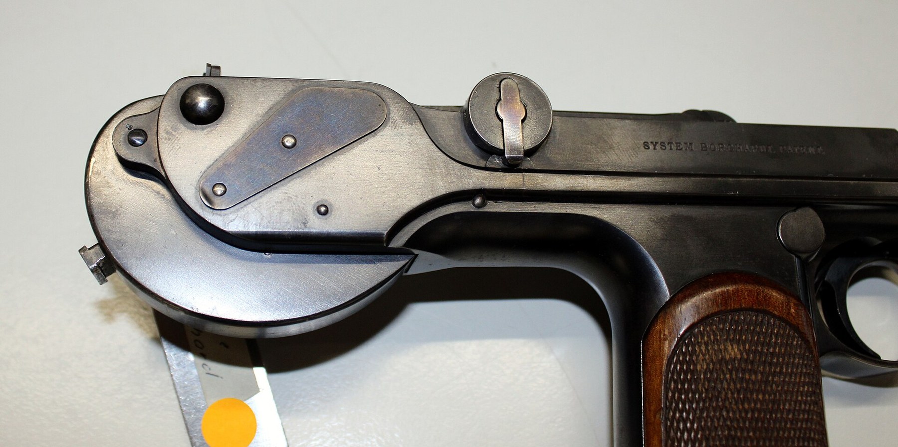 Figure 12 — The rear of a Loewe C-93, right side, showing the spring box: the round housing behind the grip, the fire-blued right cover plate over the end of the connecting pin and its slot, and the knurled to…