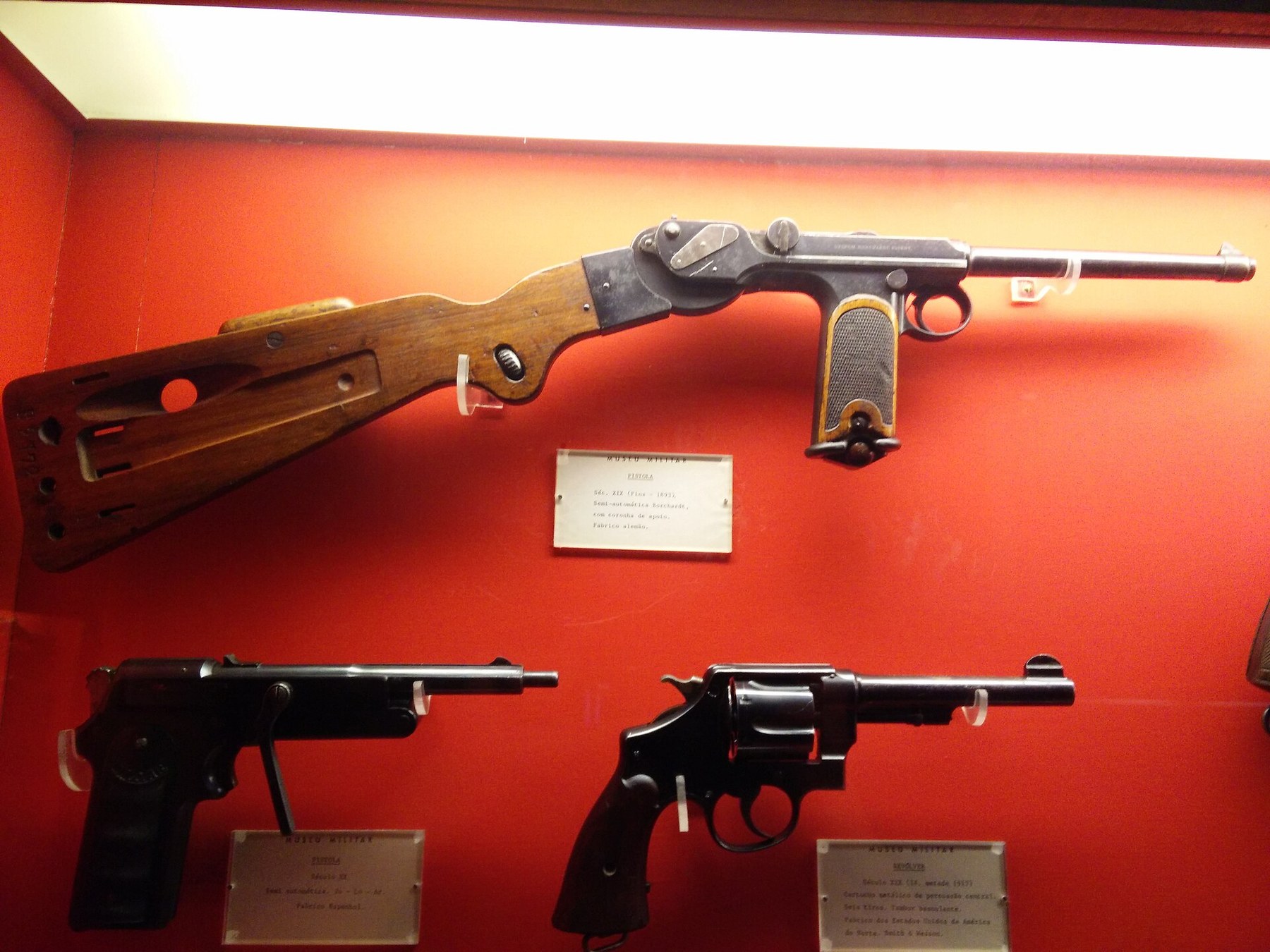 Figure 11 — A C-93 with stock in a mixed museum display, the stock's cheekpiece and the flat board clear against the red ground. The pistols below are not C-93s.