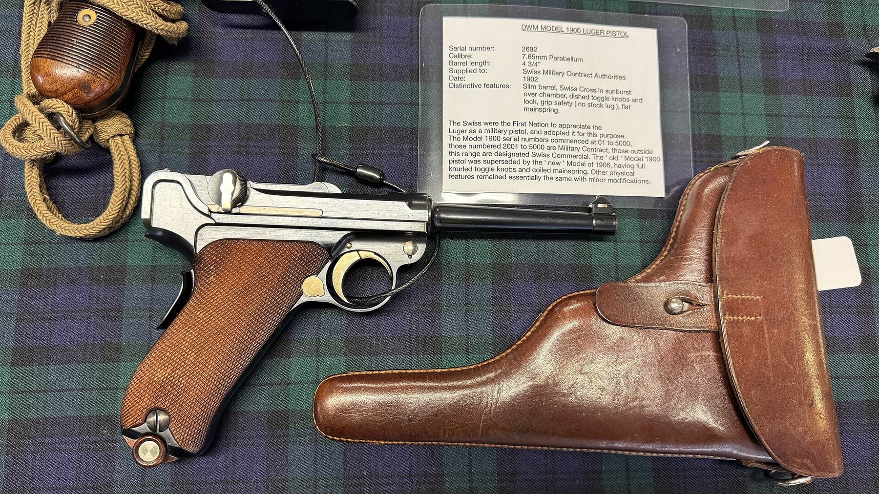 Figure 3 — A DWM Model 1900 in 7.65 mm with its holster, on display with a card naming it a Swiss military contract pistol of 1902. The card lists the features that identify the pattern: slim barrel, Swiss cr…