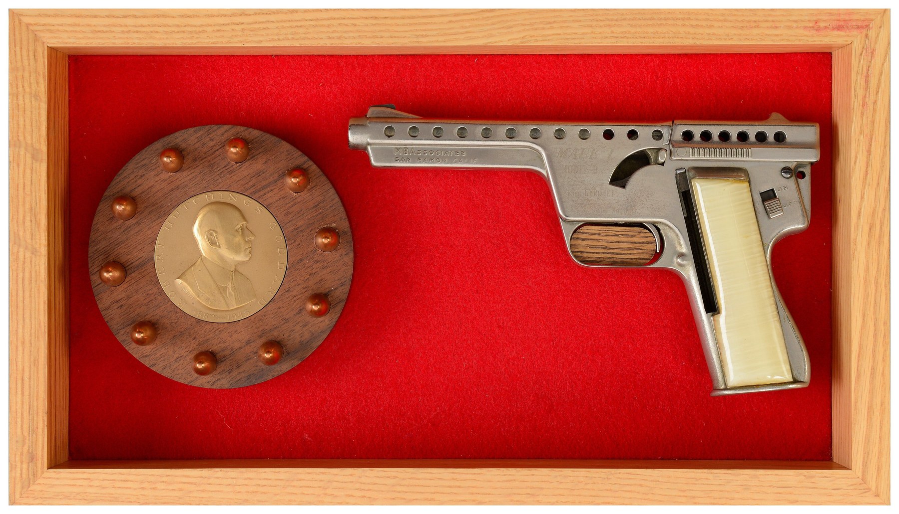Figure 5 — A nickel-finished Model B with ivory-coloured grips in an MBA presentation case, with the bronze Robert Goddard medallion surrounded by ten dummy rounds on red felt. Rock Island Auction Company pho…