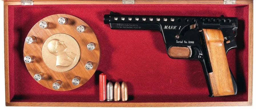 Figure 5 — An early Mark I in its walnut presentation case: the pistol, the bronze Robert Goddard medallion ringed by display rounds, and a small 13 mm red flare and inert dummies. Rock Island Auction Company…