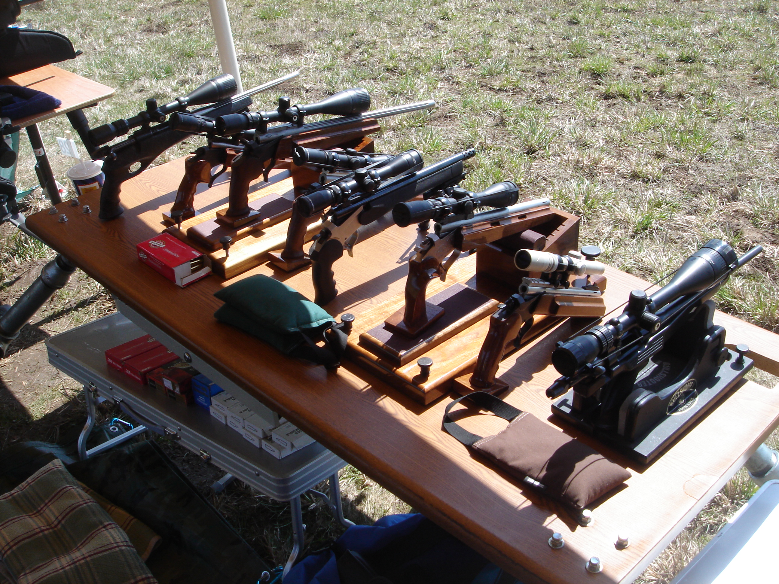 Figure 6 — Silhouette pistols on the bench at a shooting event. The long scoped bolt guns are the type the sport produced; several are XP-100 actions in aftermarket stocks. Photo: File:Metallic Silhouette Pis…