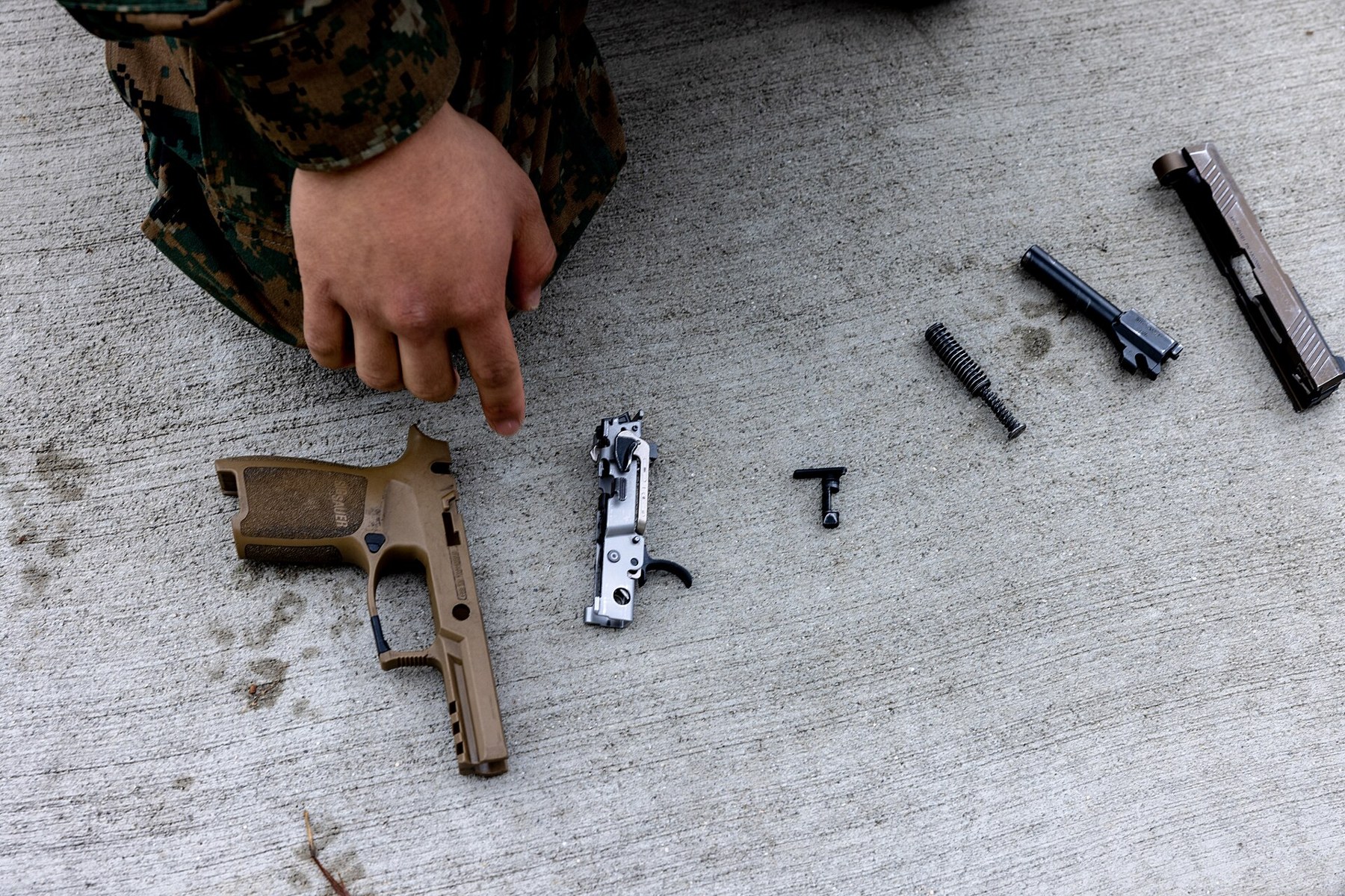 Figure 2 — An M18 detail-stripped by a Marine for a weapons-knowledge test: grip module, the stainless fire control unit with its trigger, the takedown lever, recoil spring assembly, barrel and slide. The FCU…