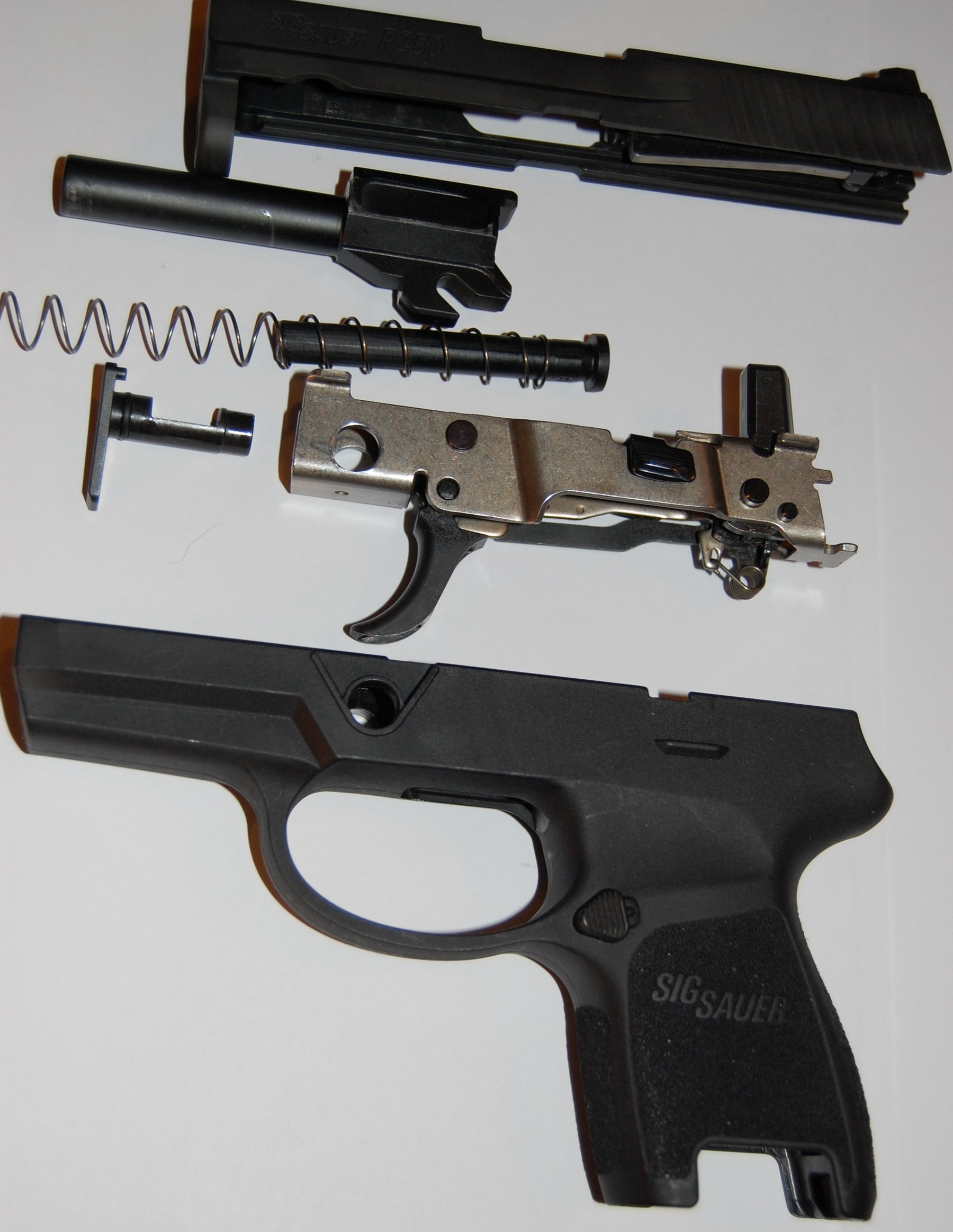 Figure 3 — A SIG P250 disassembled: slide, barrel, recoil spring, the removable fire control assembly, and the grip module. The P320 kept the removable chassis and replaced the P250's hammer with a striker.