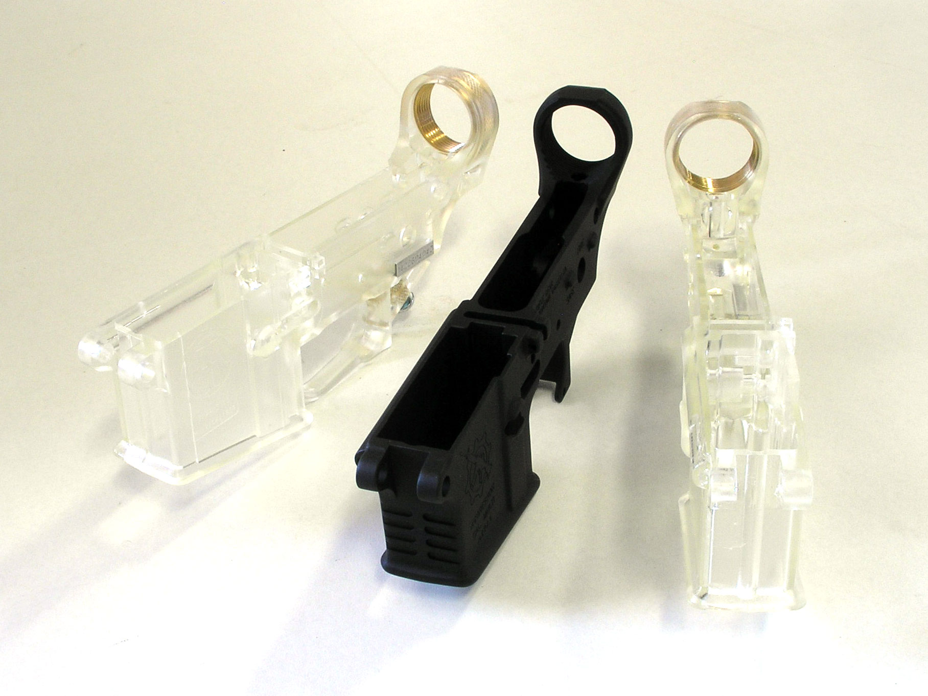 Figure 3 — Two clear polycarbonate lower receivers flanking a conventional forged aluminium one. The comparison is a useful reminder of how little structural work this part actually does — and of where the po…