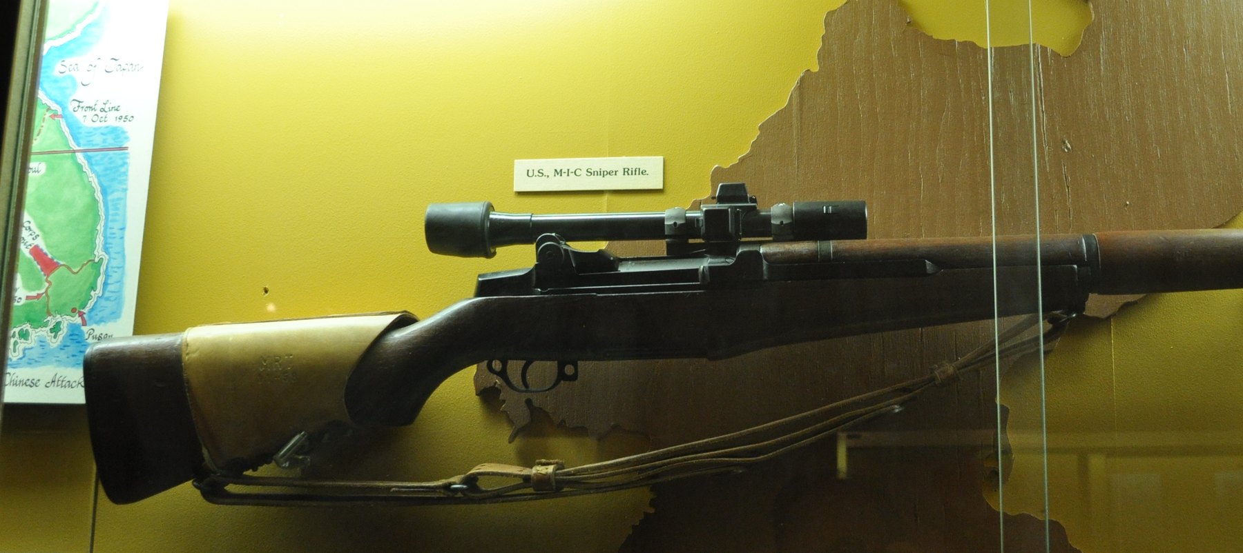Figure 4 — An M1C sniper rifle on display at the Fort Lewis Military Museum, labeled by the museum "U.S., M-I-C Sniper Rifle," with its leather cheek pad and left-side scope mount. (The photographer's own Com…