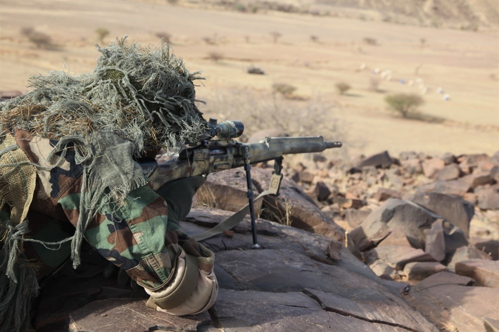 Figure 9 — A soldier in a ghillie hood with a camouflage-painted rifle during Exercise Flintlock 2013 at Nema, Mauritania, 8 March 2013. The US Army caption describes Senegalese soldiers pulling security. The…