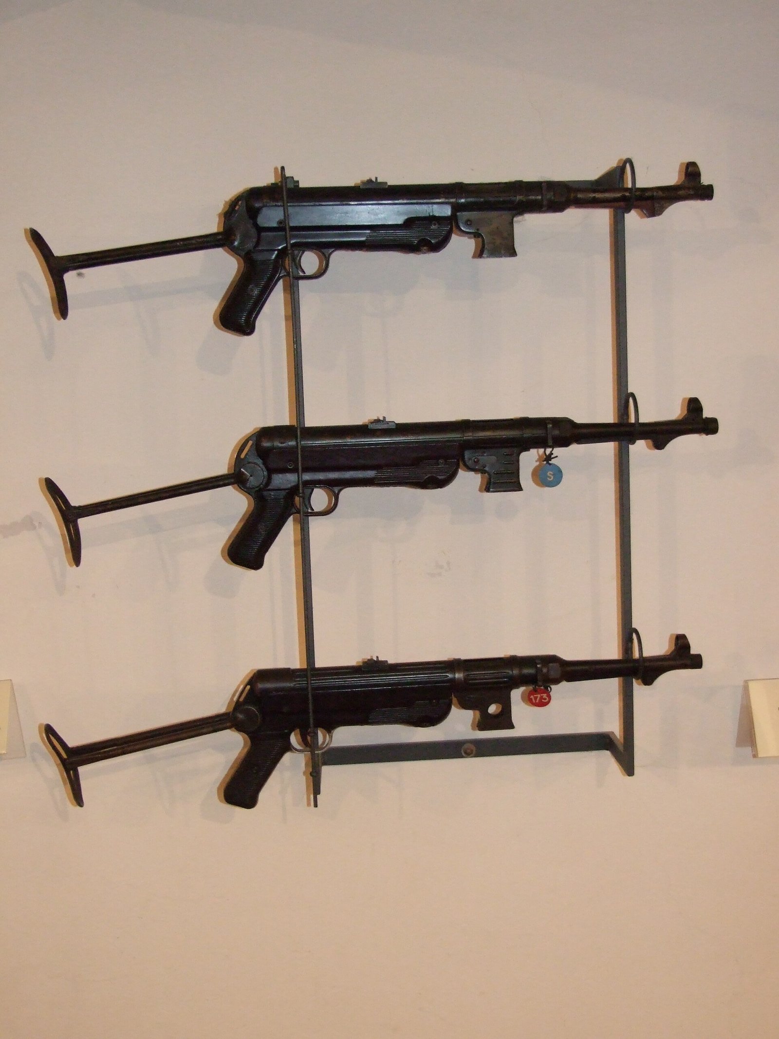 Figure 9 — An MP 38 and MP 40s together on a museum rack. The lowest weapon carries the machined longitudinal flutes of the MP 38 receiver; the receivers above it are smooth. This is the comparison to make be…