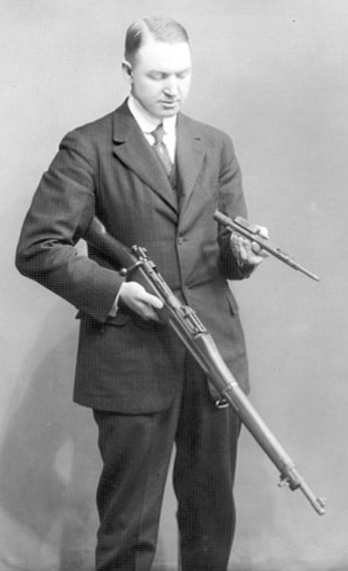 Figure 1 — John D. Pedersen holding a Springfield rifle and, in his right hand, a device that the photograph's Commons uploader identifies as the Pedersen device, c. 1917–1918. Photographer unknown; public do…