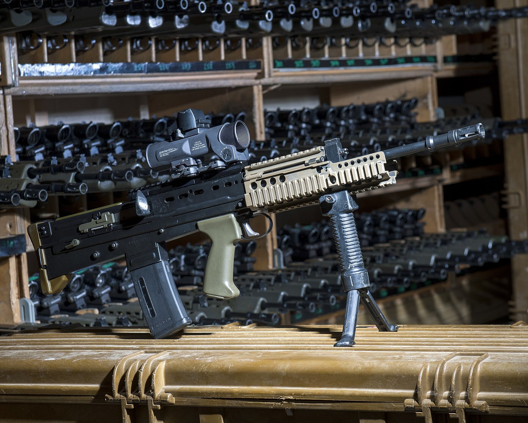 Figure 4 — An SA80-A2 in a unit armoury rack, with an optical sight, rail fore-end and Grip Pod; the racked rifles behind it. MOD photograph by Stuart Hill, Open Government Licence v3.0.