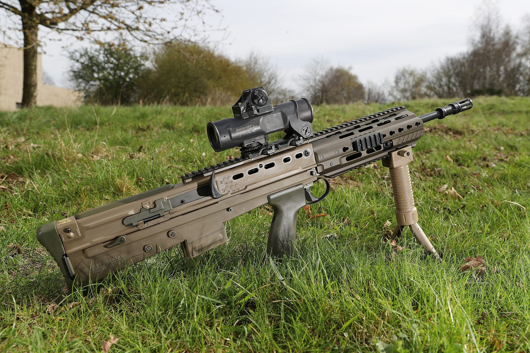 Figure 9 — A Flat Dark Earth L85A3 lying on grass with its bipod-grip deployed and an optical sight, 2018. The continuous top rail and the H&K fore-end are visible. MOD photograph by Graeme Main, Open Governm…