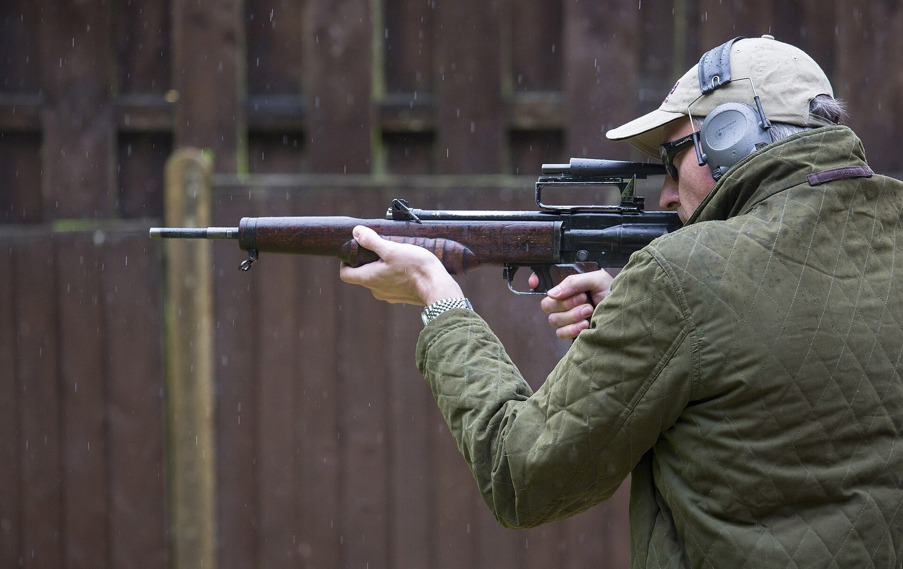 Figure 4 — A civilian in ear defenders fires the same EM-2 at the 2016 Ministry of Defence event, showing the rifle's high optical sight in its carrying handle and the magazine behind the grip. MOD photograph…