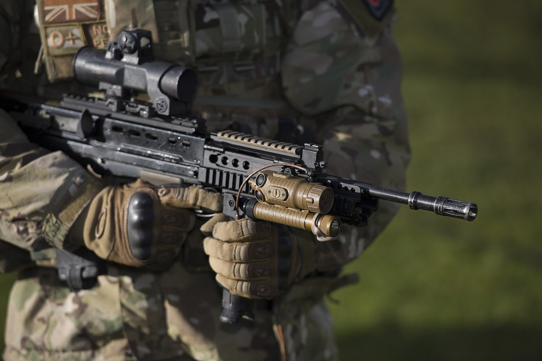 Figure 8 — An upgraded Laser Light Module Mk 3 on the side rail of an SA80-A2, held by a soldier; a switch cable loops back toward the firer's support hand, and an Elcan with a small reflex sight is on the bo…