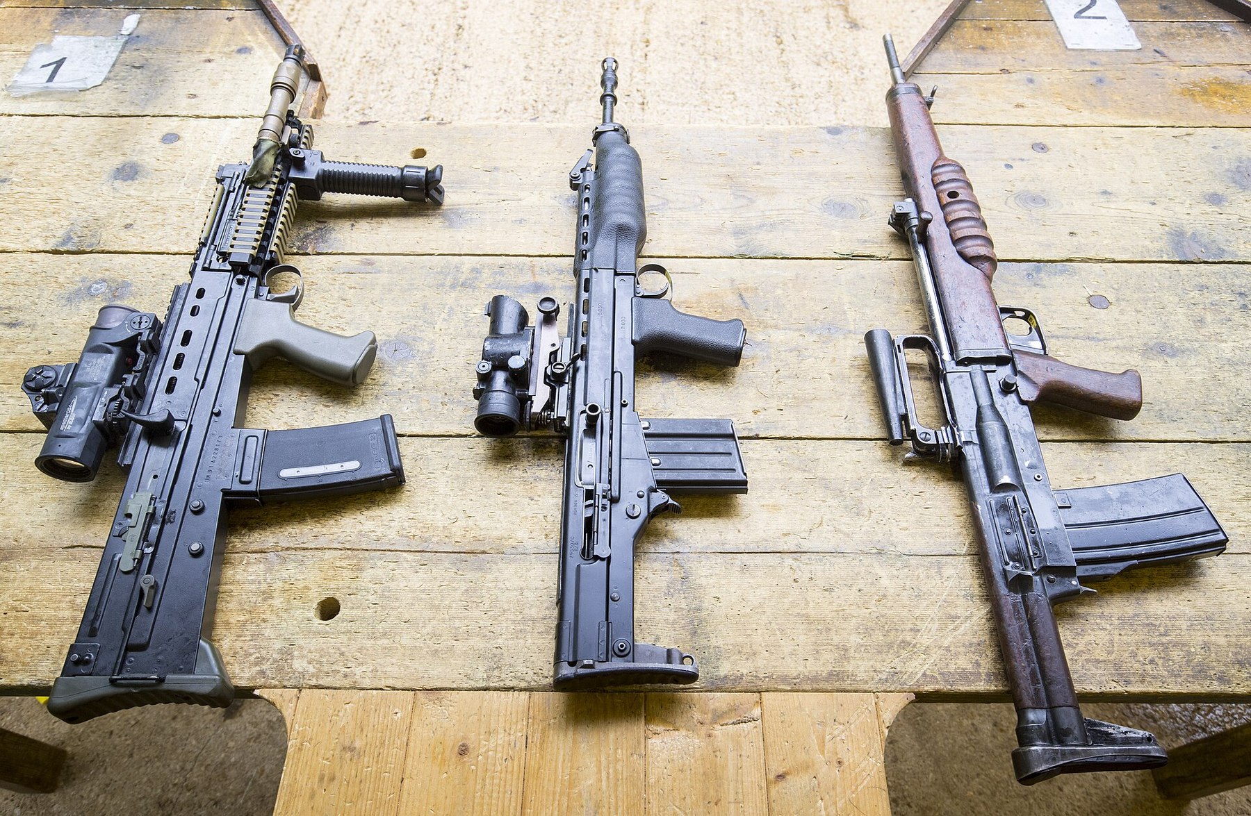 Figure 12 — Three British rifles on a wooden bench, left to right: an SA80-A2 with rail fore-end and optical sight, an XL60-series prototype with its optical sight removed and placed beside it, and the wooden-…