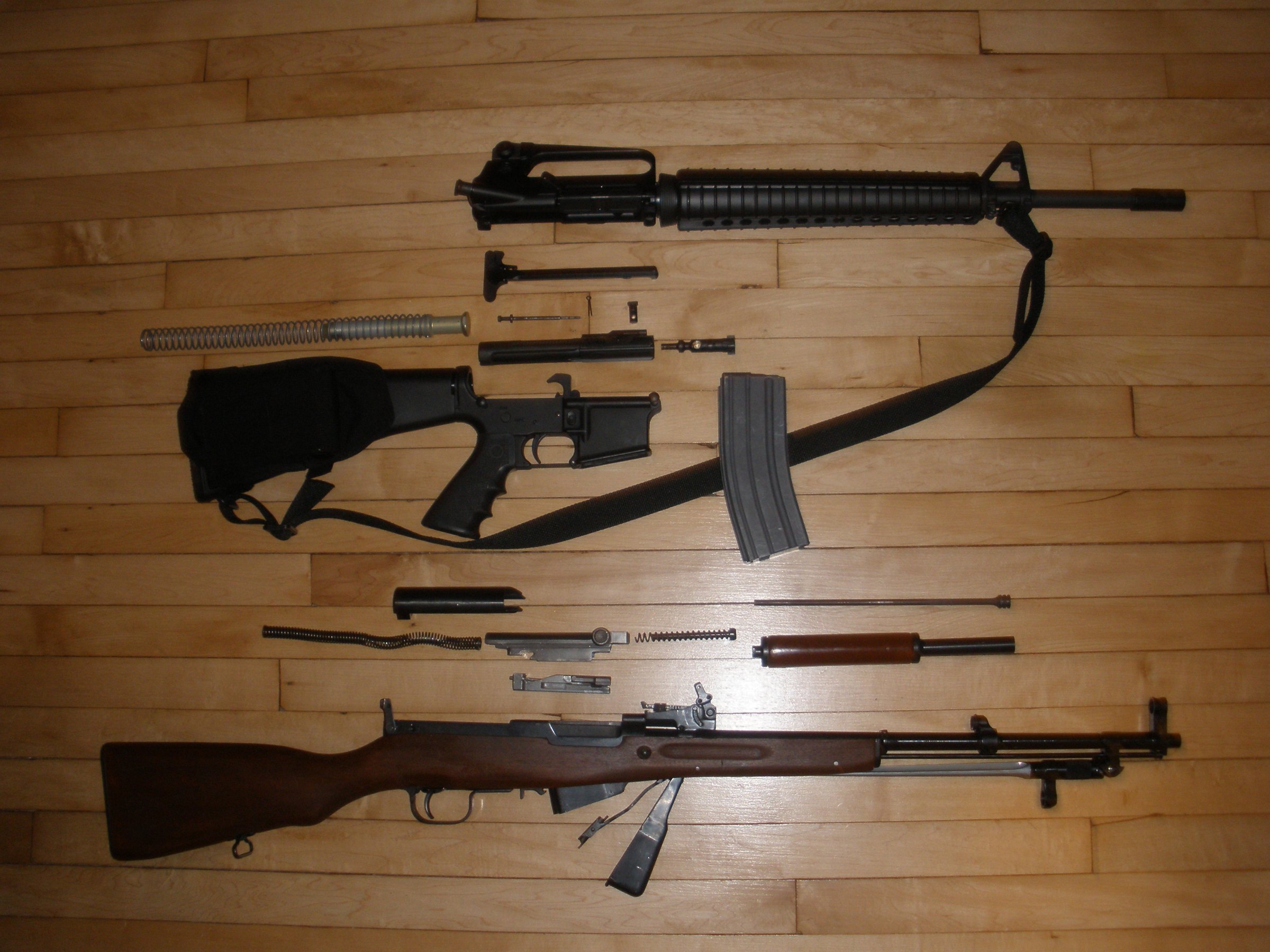 Figure 8 — An SKS and an AR-15 field stripped side by side. Six groups against roughly a dozen, no tools against no tools, and the SKS needs nothing that is not already on the rifle.