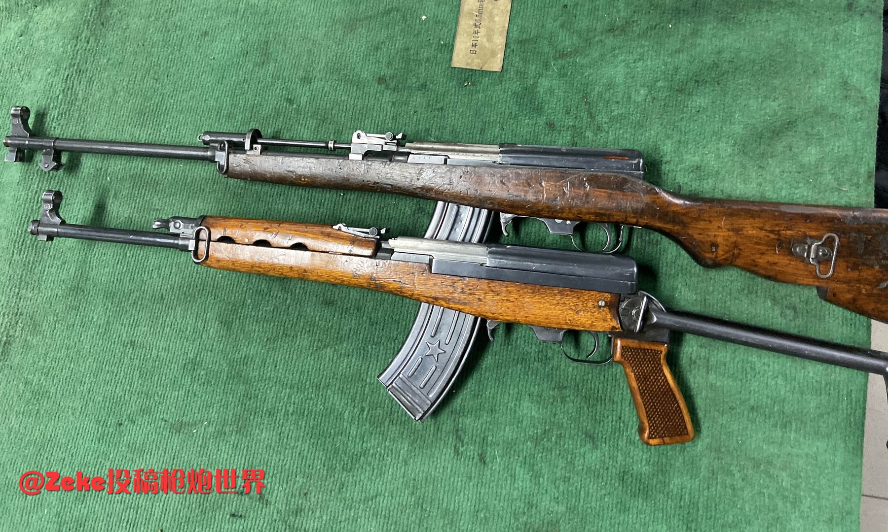 Figure 5 — A standard Type 63 above and the airborne folding-stock sample below, photographed together on a collector's mat, muzzles left. The sample has a separate wooden pistol grip and an underfolding stee…