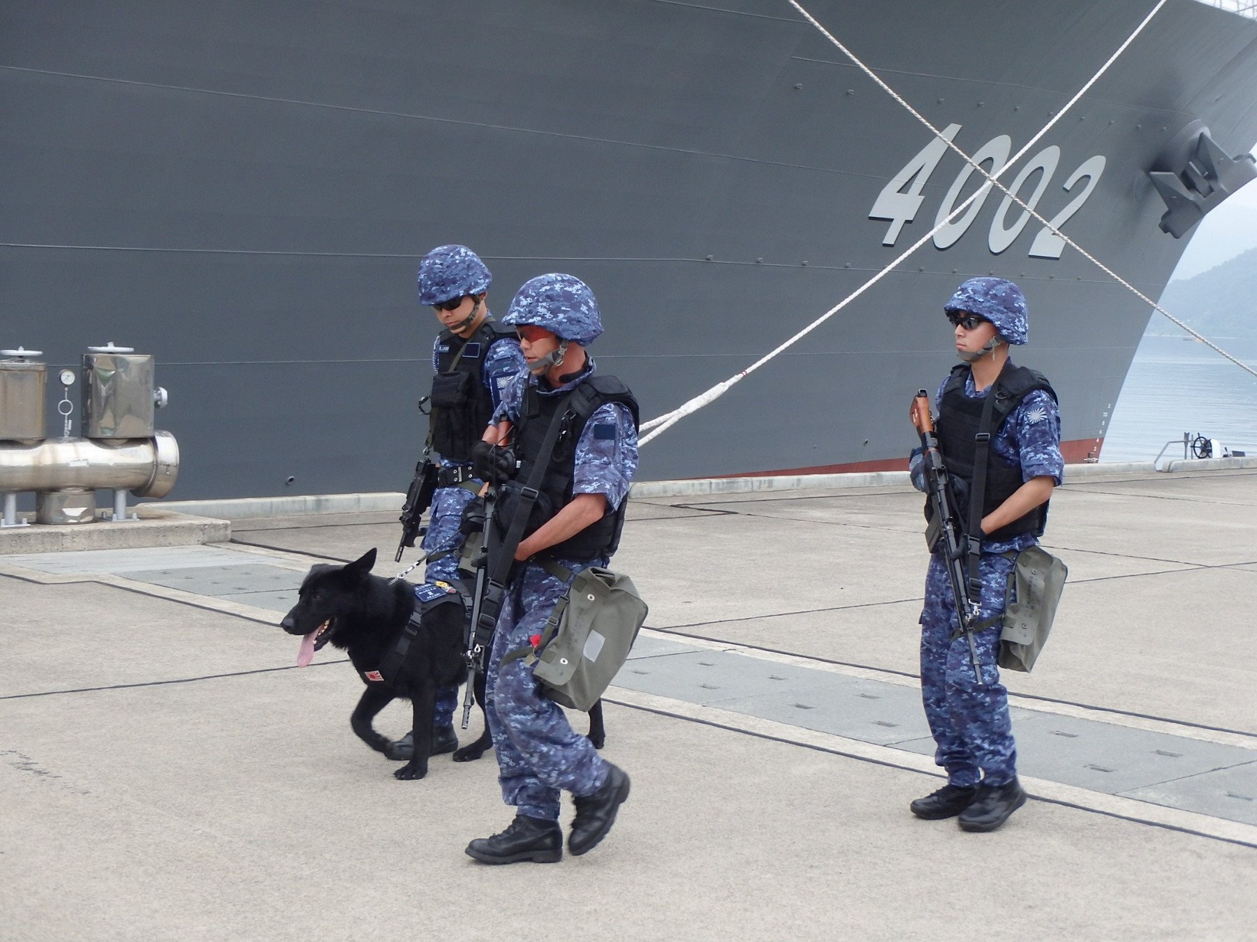Figure 3 — Members of the Maizuru District security unit guard the quay at Maizuru with a working dog, July 2019. The JMSDF's own caption: "武器としては、64式小銃（一般隊員）、9ｍｍ機関けん銃（警備犬ハンドラー）を装備" (armed with the Type 64 ri…