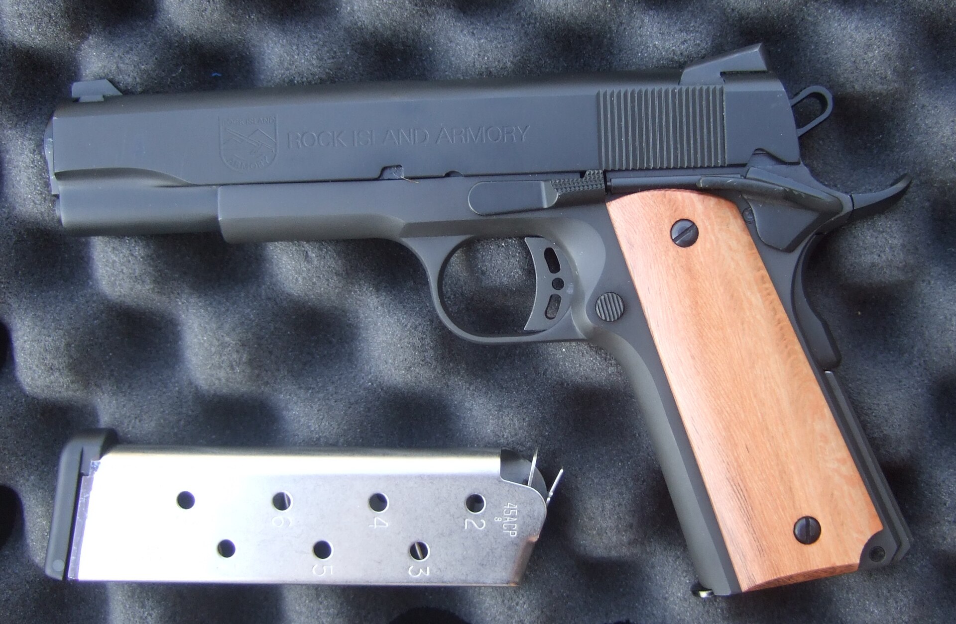 Figure 3 — A Rock Island Armory "Tactical" 1911 pistol photographed in 2009 in its foam case, with wooden grip panels, a skeletonised hammer and a spare stainless magazine. The 1911 is the product the Rock Is…