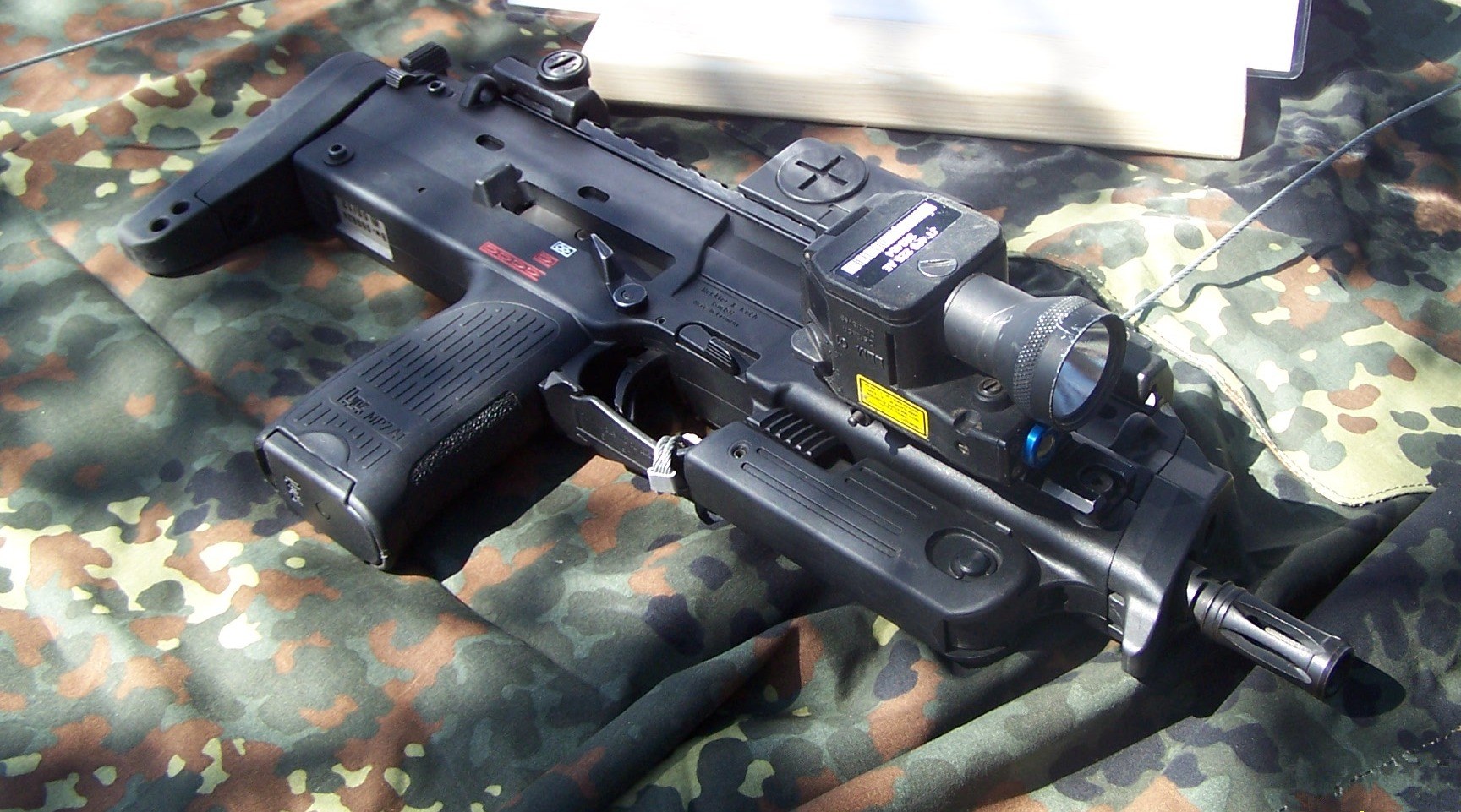 Figure 9 — An MP7 on a Bundeswehr display table, right side, with the stock extended, the folding grip folded, a reflex sight on the top rail and a laser-light module on the right-side rail. The selector leve…