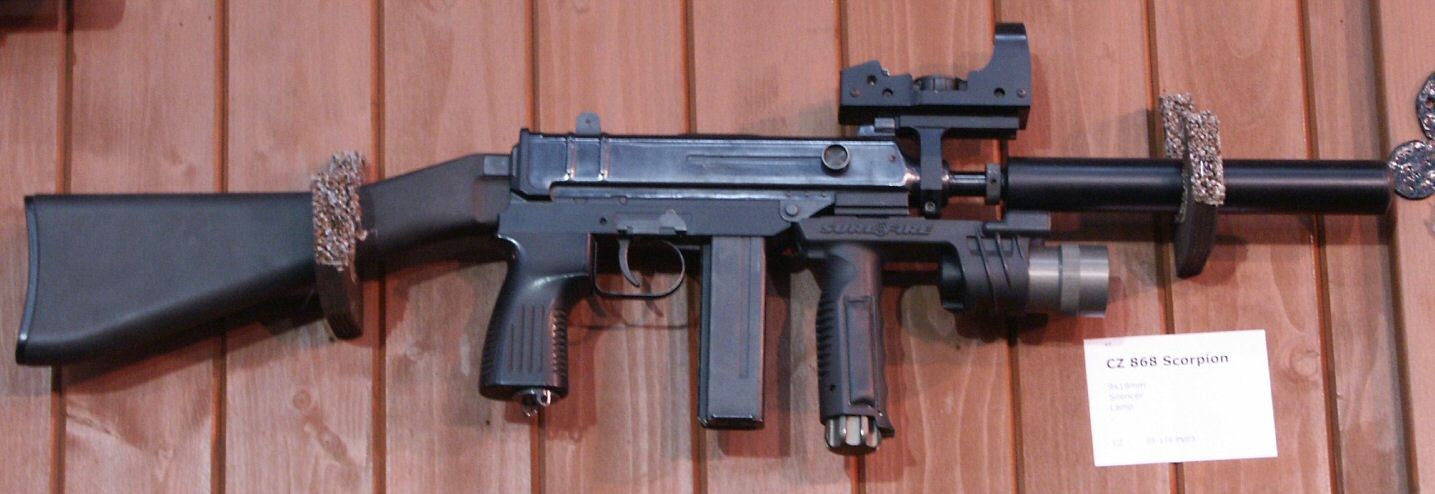 Figure 7 — A CZ 868 at a trade show, with fixed butt, vertical foregrip, top rail and red-dot sight, and suppressor; display card "CZ 868 Scorpion". Photograph by Pibwl, CC BY-SA 3.0, via Wikimedia Commons.