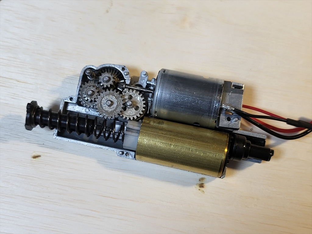 Figure 7 — The Tokyo Marui electric MP7A1 mechbox opened. The motor (top right, with red and black leads) lies parallel to the brass cylinder (bottom), with the gear train and the spring and guide at the left…