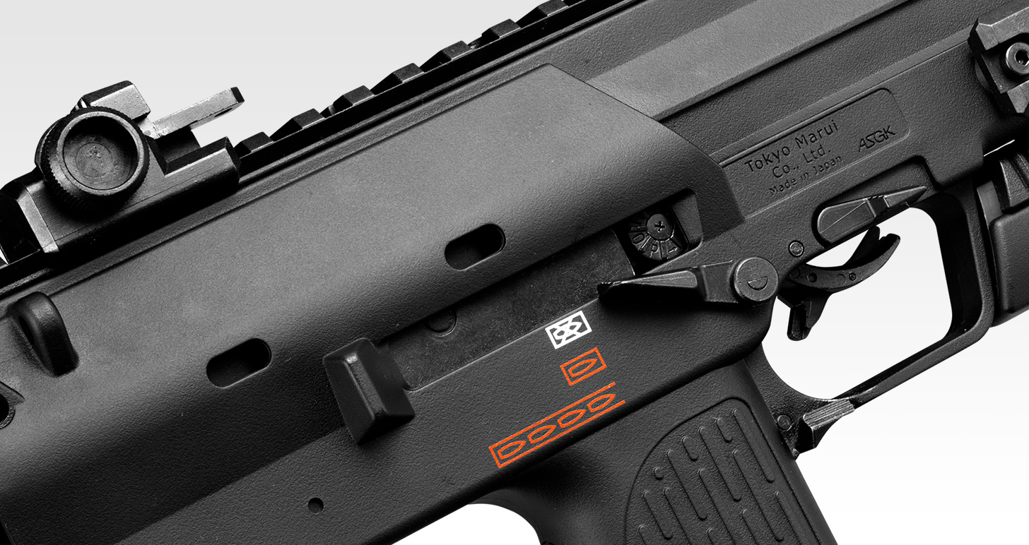 Figure 3 — The left side of a Tokyo Marui MP7A1 gas gun above the trigger. The receiver plate reads Tokyo Marui Co., Ltd., Made in Japan, ASGK, and carries no H&K trademark. The red and white symbols are the …