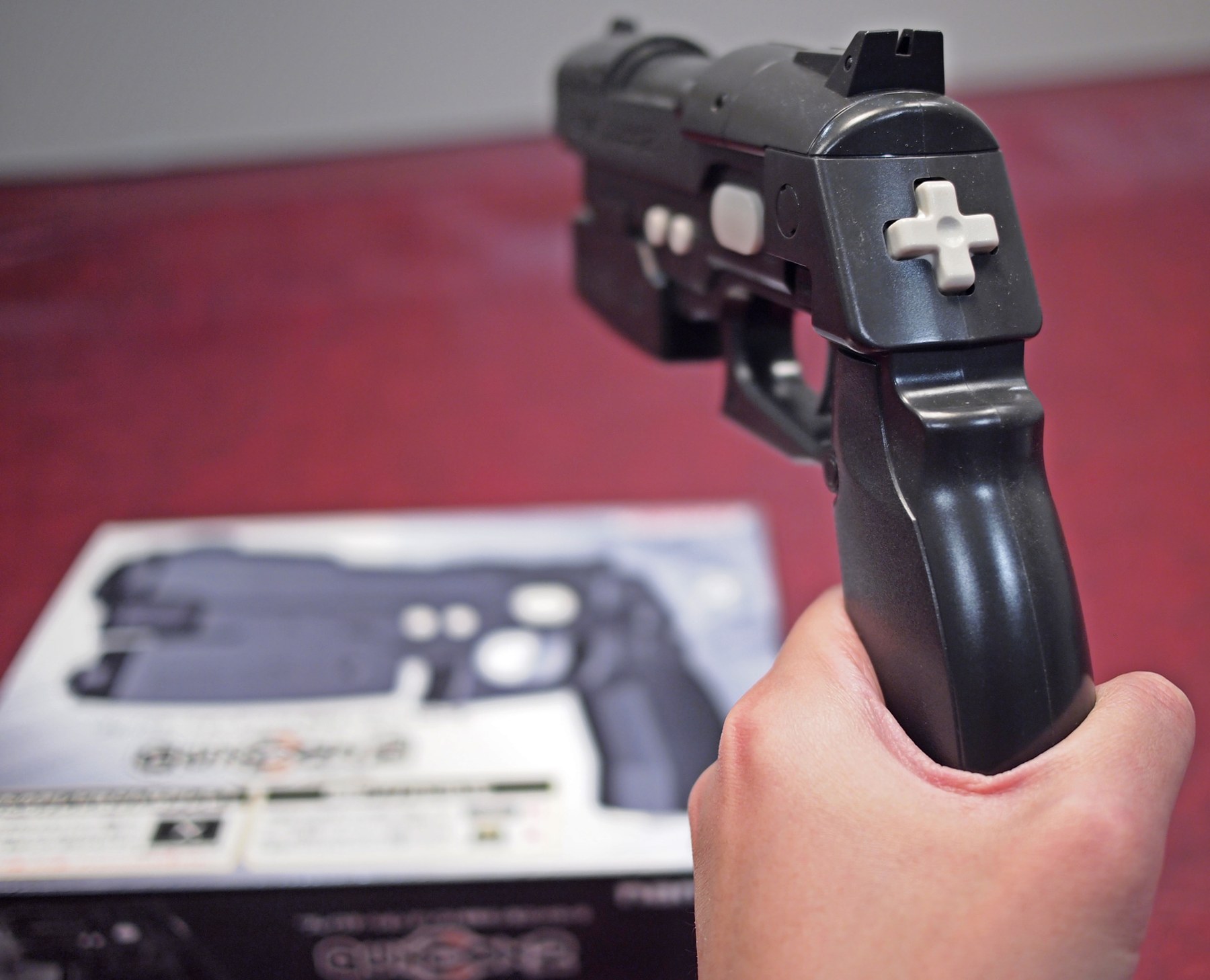 Figure 5 — The Japanese GunCon 2 in black, held in the hand in front of its box. On the rear of the slide is the D-pad Namco's manual calls the "Thumbcon". Photo: Chinpokomon5, Wikimedia Commons, CC BY-SA 3.0.