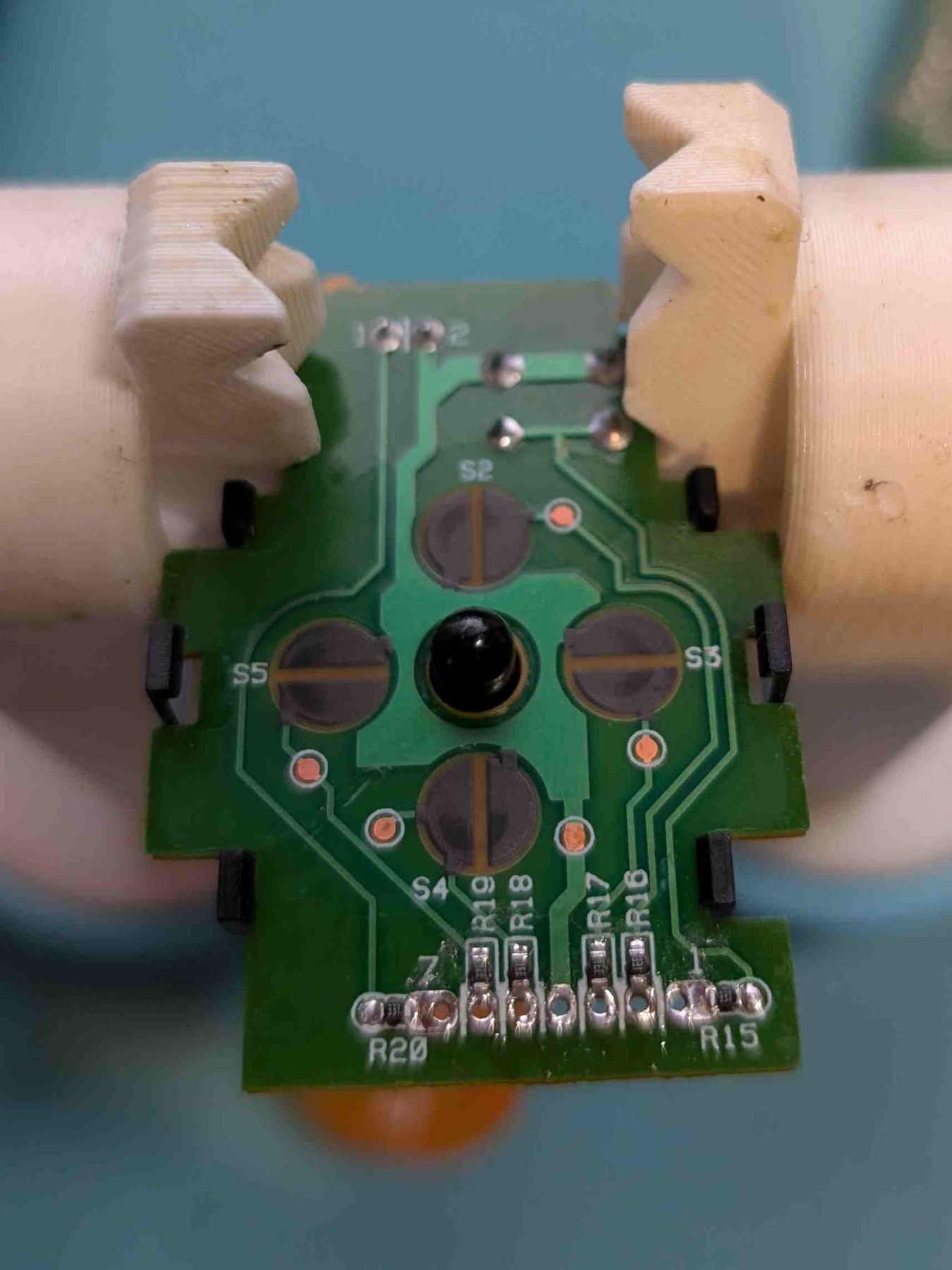 Figure 3 — The D-pad and trigger board with its ribbon desoldered: the four D-pad contacts around the centre, the seven-way row of vias at the bottom edge, and the board held between printed jaws. Photo: The …