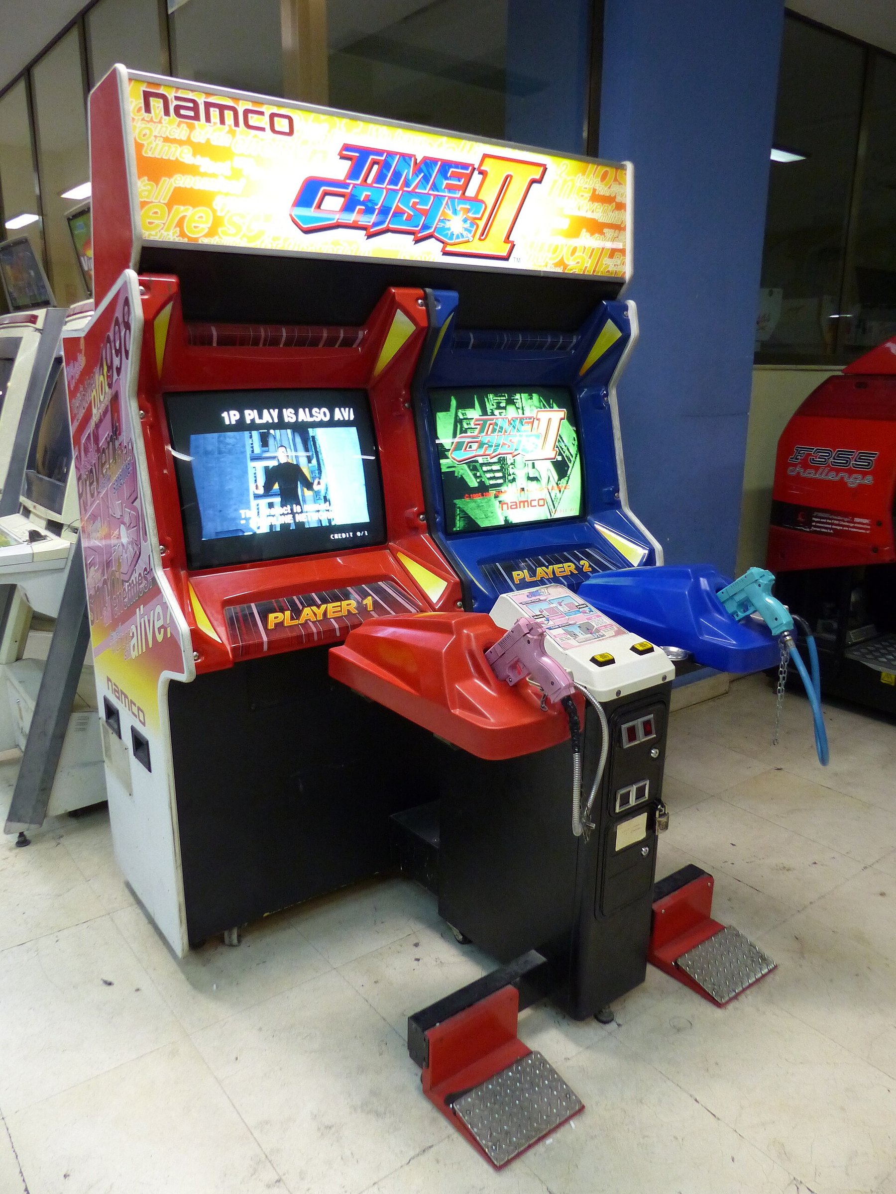 Figure 2 — A Time Crisis II twin cabinet in Spain, 2017. The guns hang on holsters in the red and blue consoles; the pedals at floor level are the "take cover" control that defines the series. The recoil supp…
