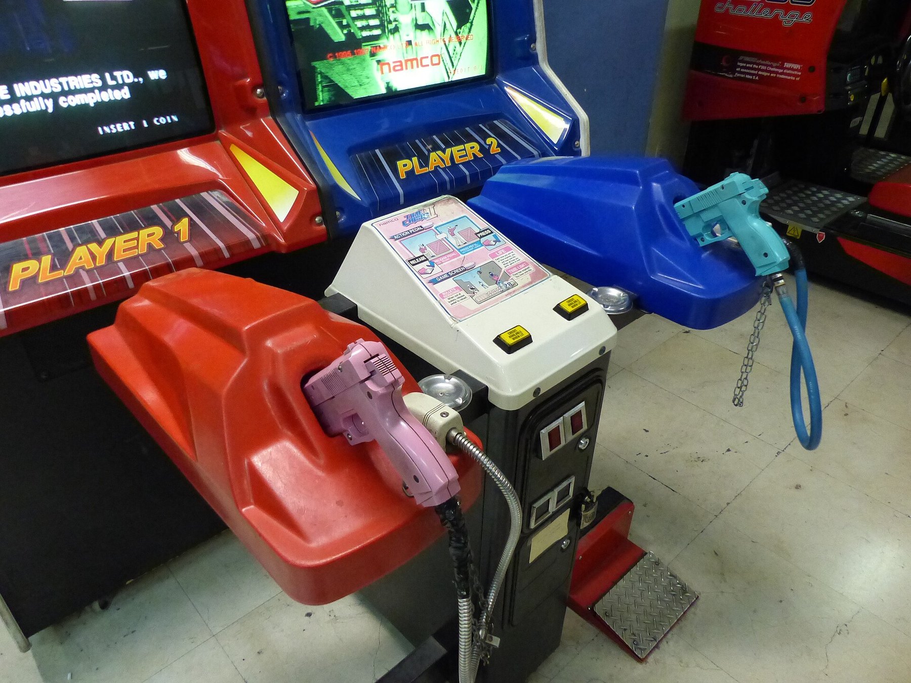 Figure 1 — Two Time Crisis II guns on their cabinet holsters, pink for player one and blue for player two, photographed in Spain in 2017. The upper part of each gun is a separate slide; a solenoid inside the …